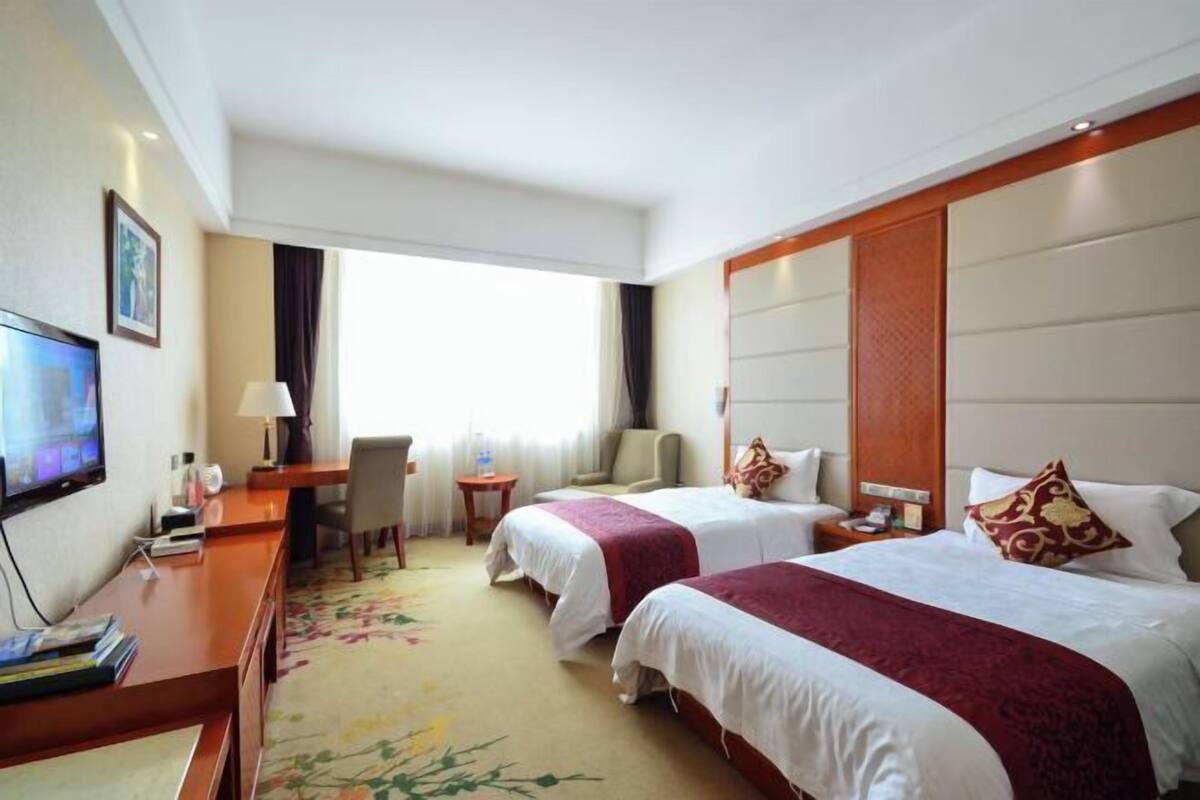 Habitación de hotel con dos camas, un escritorio con televisor, una silla, una mesita y una ventana con cortinas.
