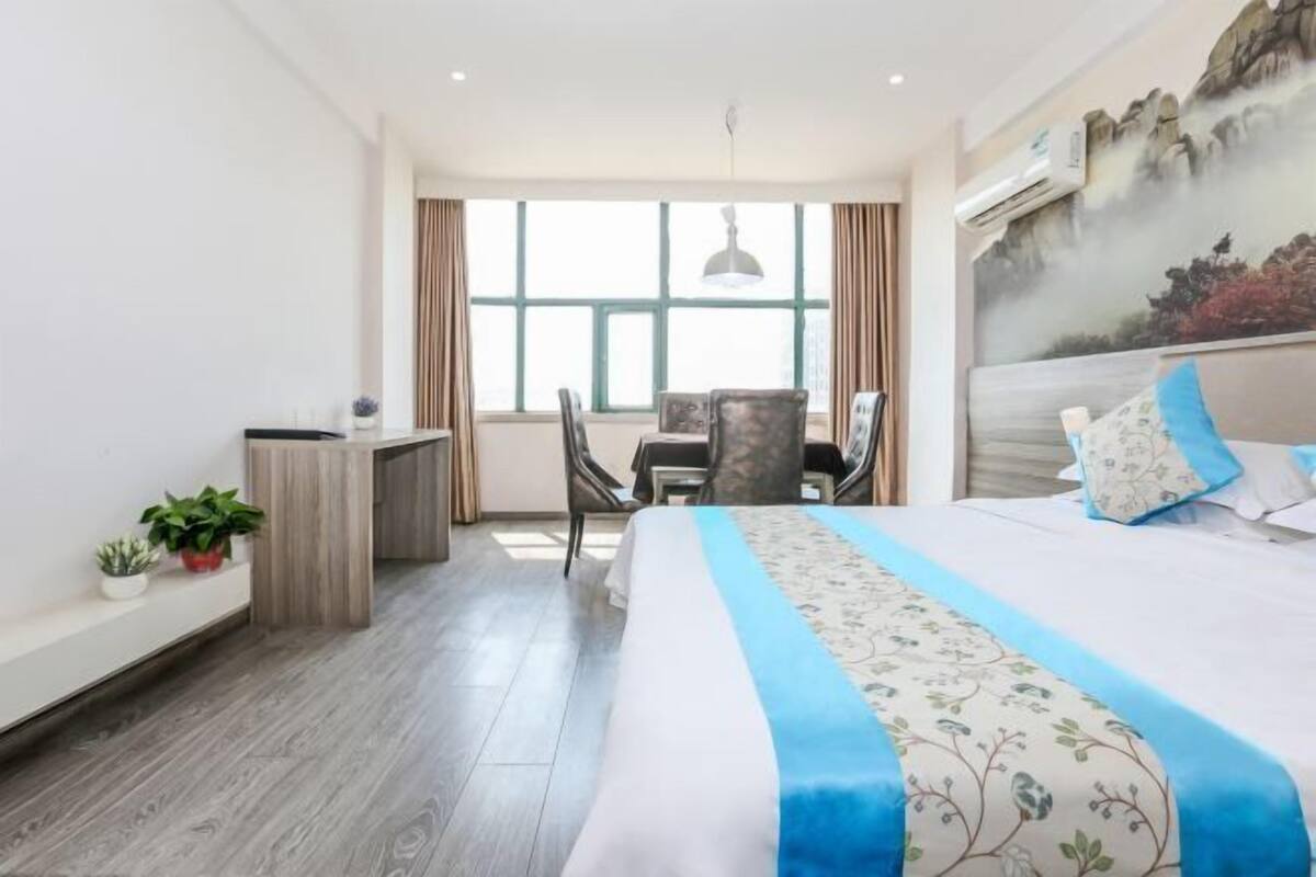 Ein Hotelzimmer mit Bett, Schreibtisch und Stuhl. Ein Fenster mit Vorhängen, ein Bild an der Wand und ein Blumentopf auf dem Schreibtisch.