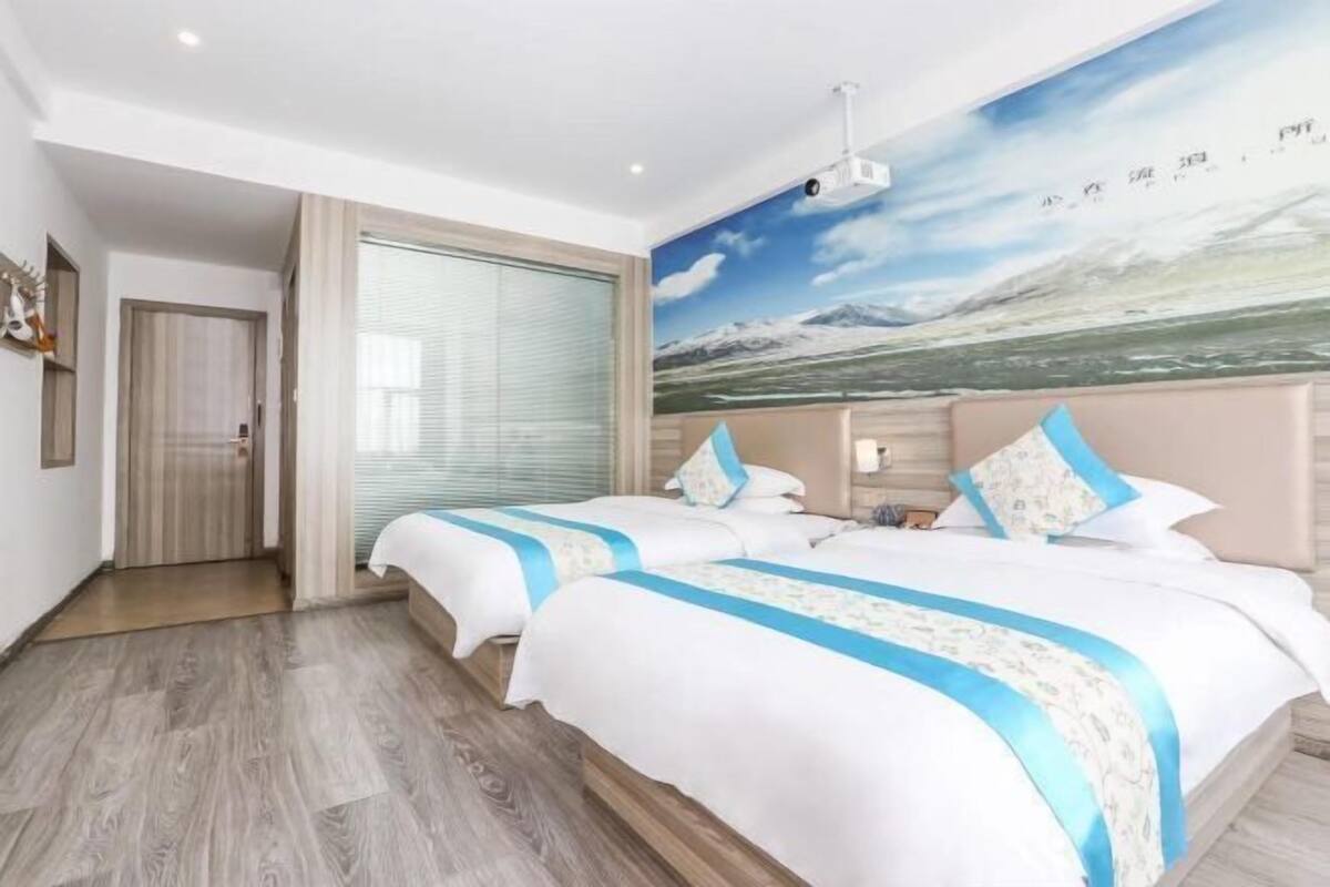 Ein Hotelzimmer mit zwei Betten, einem großen Fenster mit Jalousien, einem Deckenprojektor und einem Wandbild einer Berglandschaft.