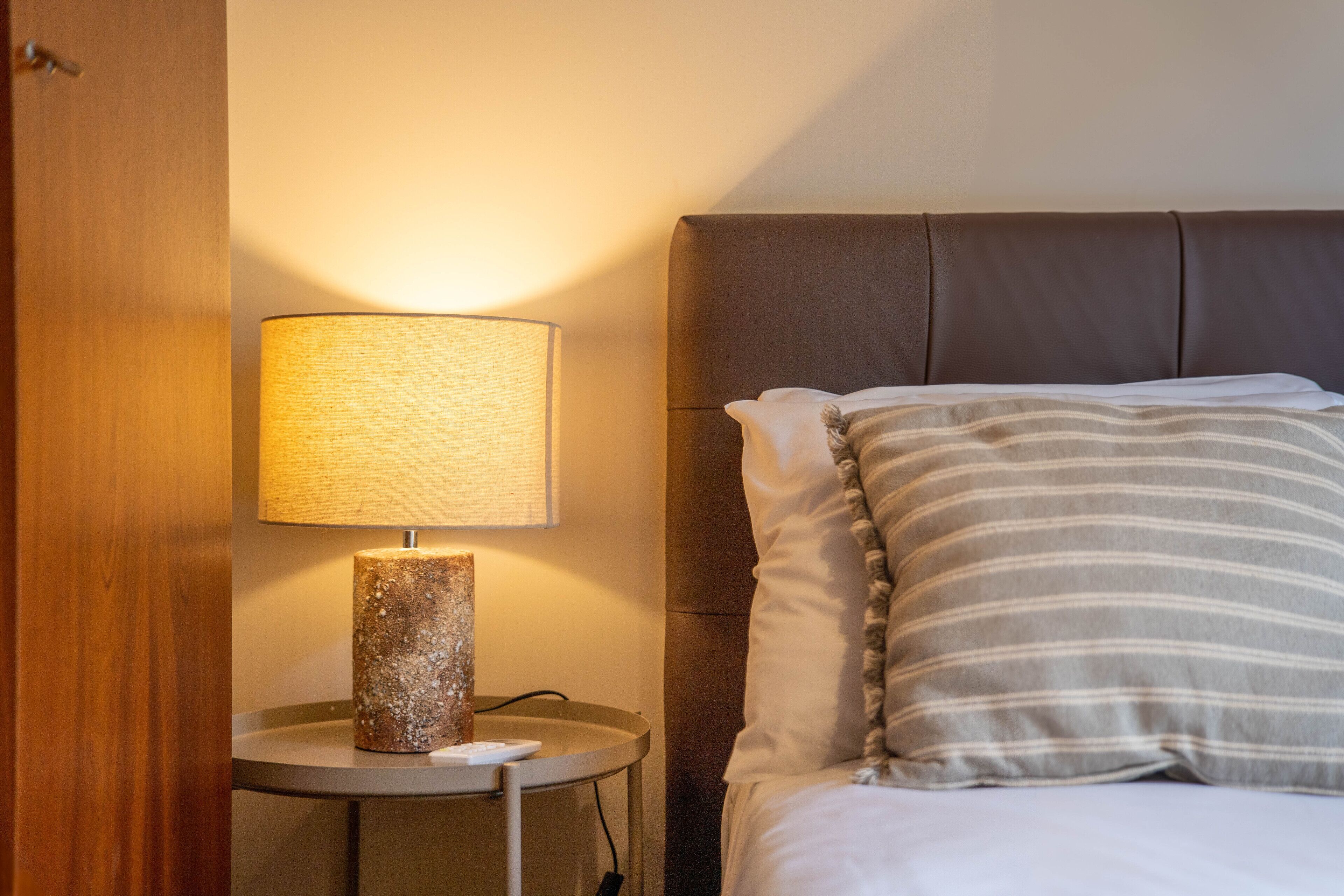 A neatly made bed with striped pillows, a round bedside table with a textured lamp, and a wooden door.