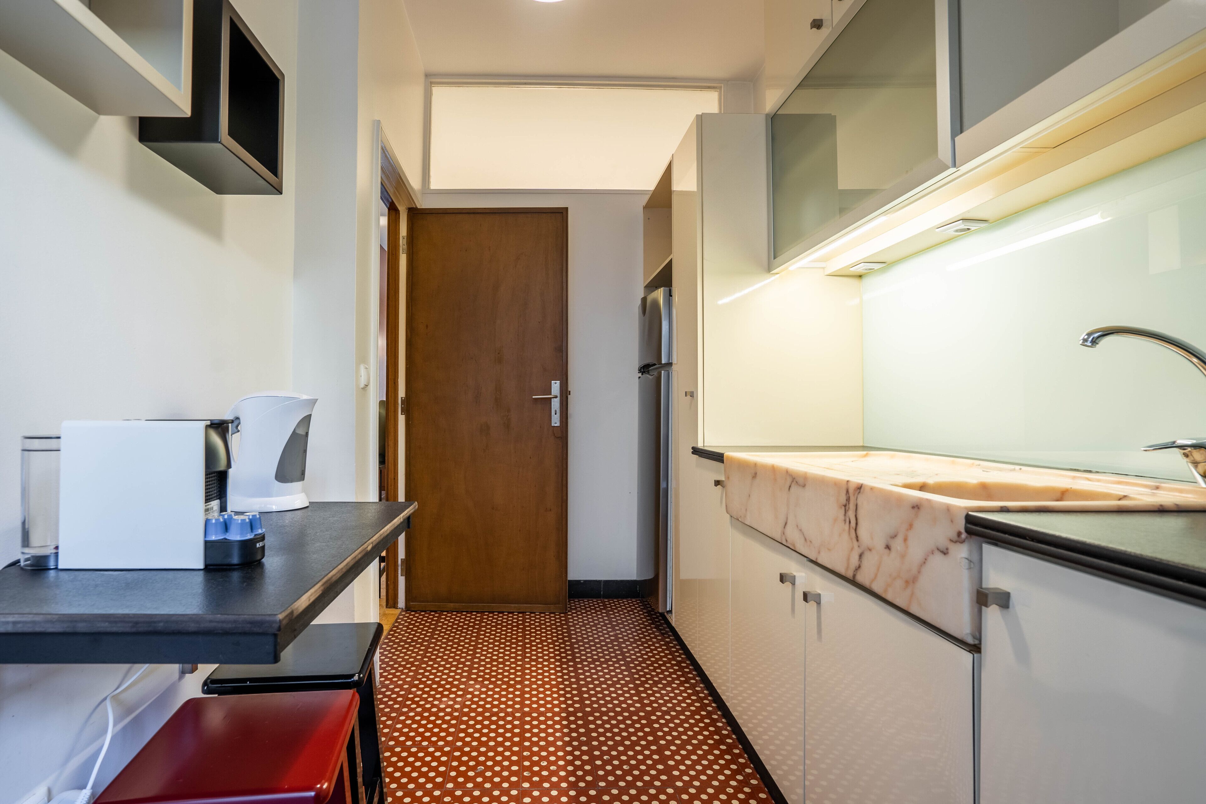 Ein kleines Hotelzimmer mit einer Küchenzeile, einer Holztür, einem gemusterten Boden und einem Waschbecken mit Marmorplatte.
