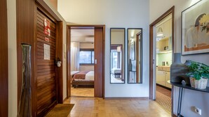 Pasillo de hotel con puertas de madera, una cama al fondo, un espejo y una planta sobre una mesa.