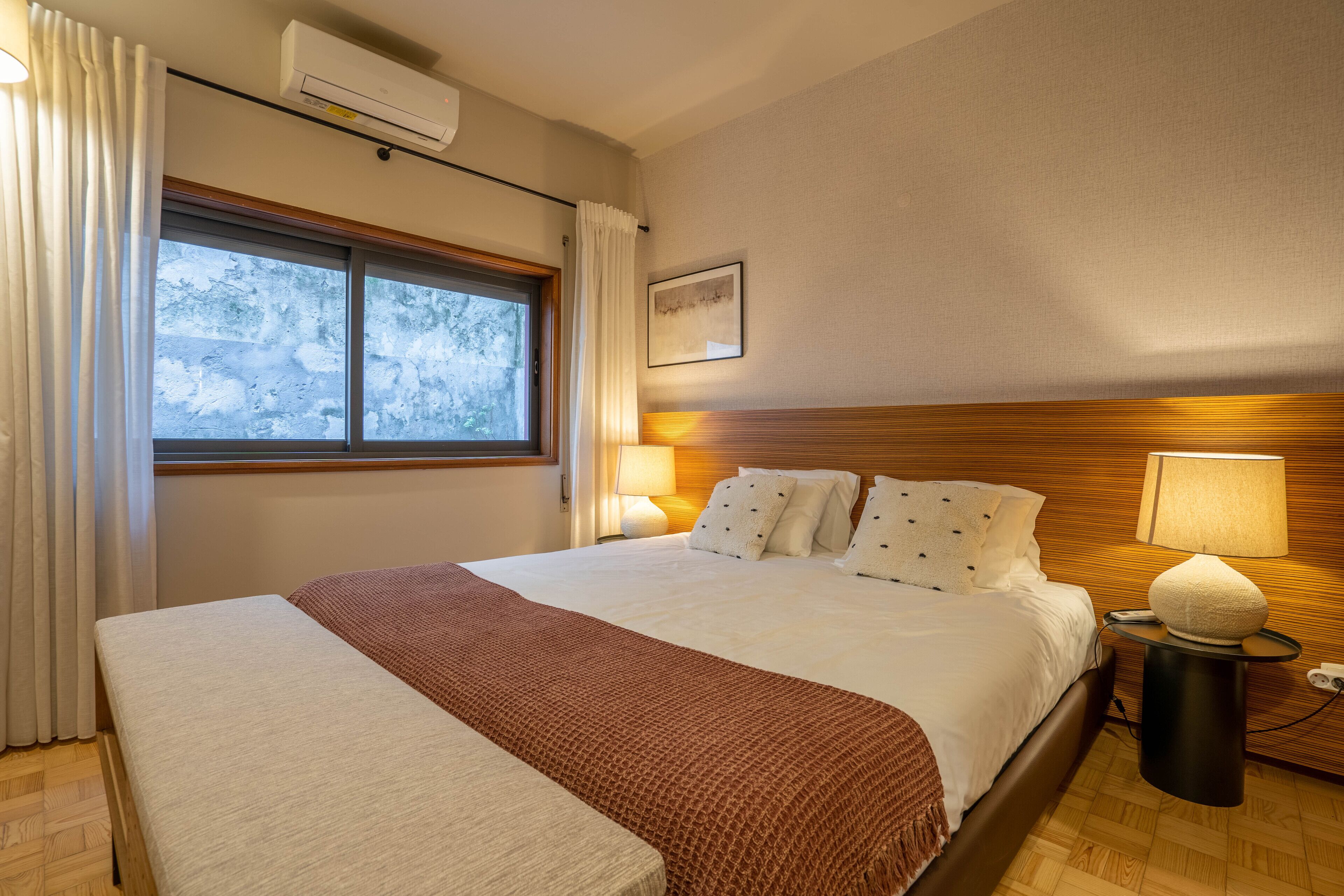 A hotel room with a large bed, a bedside table with a lamp, a window with curtains, and an air conditioning unit.