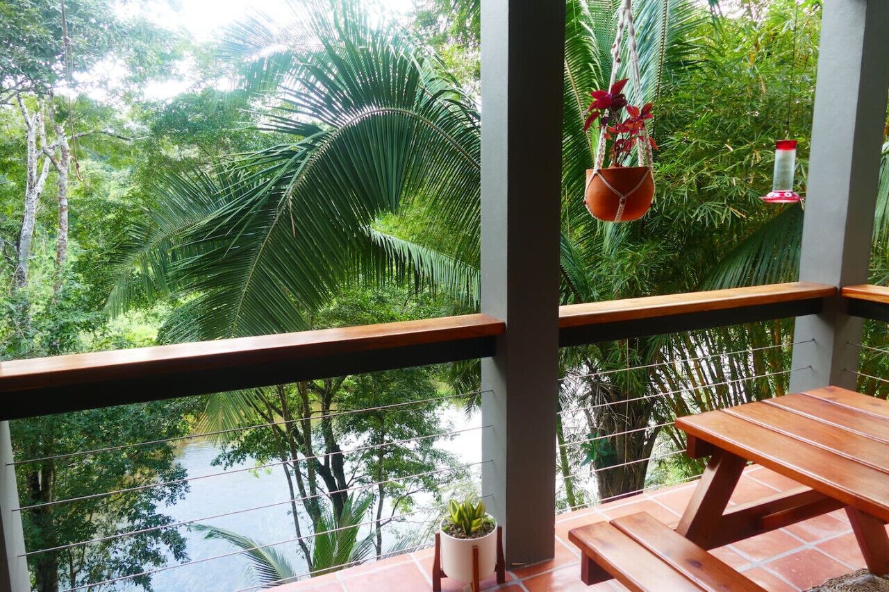 A balcony with a wooden table and chairs, overlooking a lush green landscape with palm trees and a body of water.