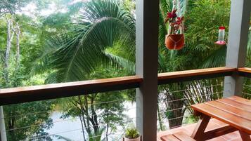 A balcony with a wooden table and chairs, overlooking a lush green landscape with palm trees and a body of water.