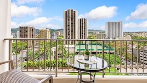 A balcony with a table and chairs, overlooking a cityscape with tall buildings and a tennis court.