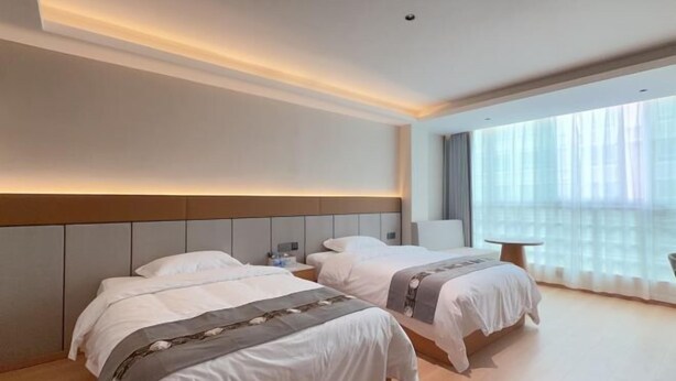Quarto de hotel com duas camas, mesa de cabeceira, cadeira e uma janela grande com cortinas translúcidas.