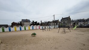 En sandstrand med färgglada strandhytter, en lekplats med gungor och en rutschkana, och hus i bakgrunden.