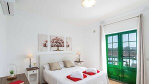 Une chambre d’hôtel avec un lit, des tables de chevet, une commode, une fenêtre avec un cadre vert et des œuvres d’art accrochées au mur.