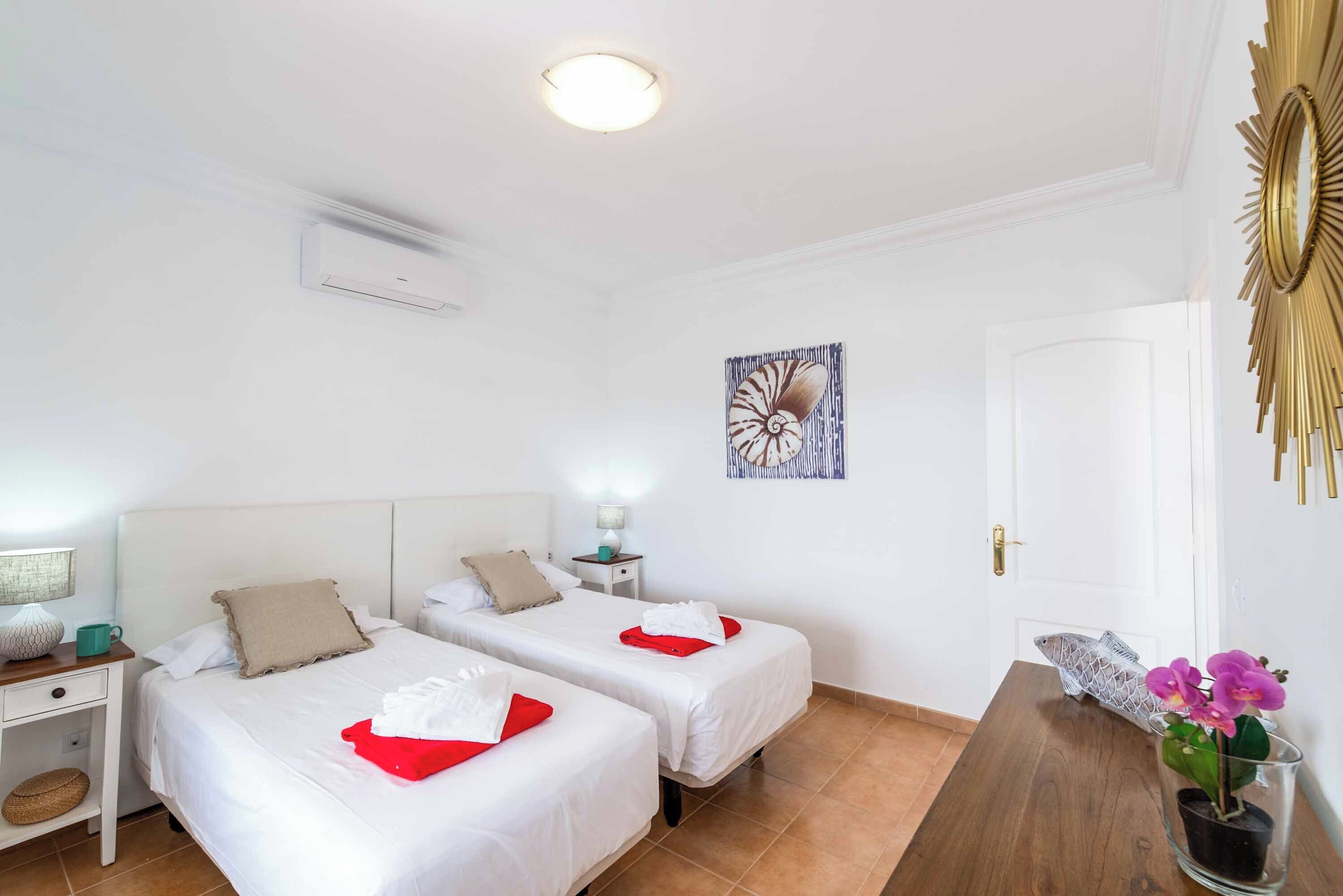 A hotel room with two beds, a wooden nightstand, a lamp, a wall art piece, and a door with a decorative mirror.