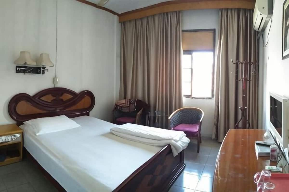 A hotel room with a bed, a desk, a chair, a window with curtains, and an air conditioning unit.