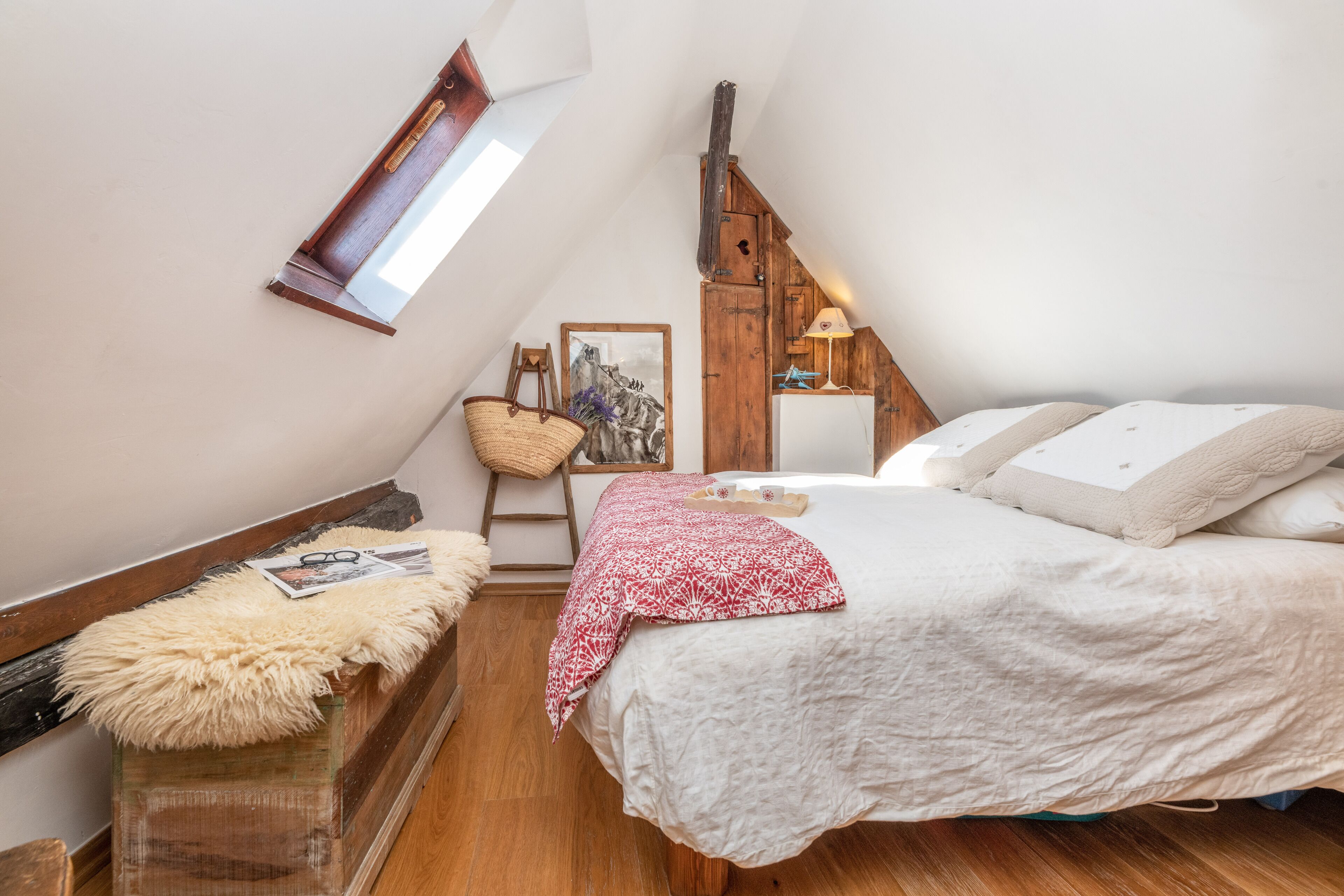 A cozy bedroom with a sloped ceiling, a wooden ladder, a basket, a framed picture, and a bed with white bedding.