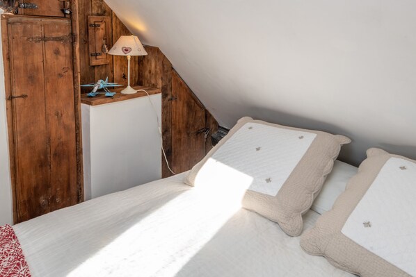 A bedroom with a sloped ceiling, a wooden wardrobe, a bedside lamp, and a bed with a quilted cover.