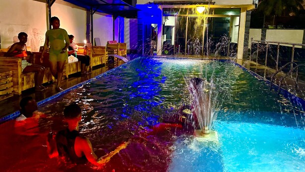 A swimming pool with a water fountain, surrounded by wooden benches and a covered area. - Northwold Hotel-Sowutuom Accra (Accra)