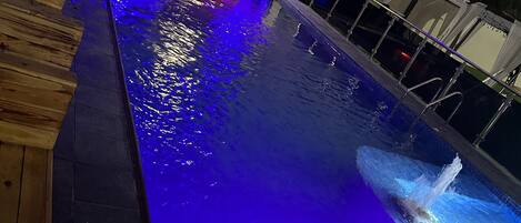 Una piscina con una cubierta de madera en un lado, un elemento decorativo de agua y un edificio con iluminación azul al fondo.