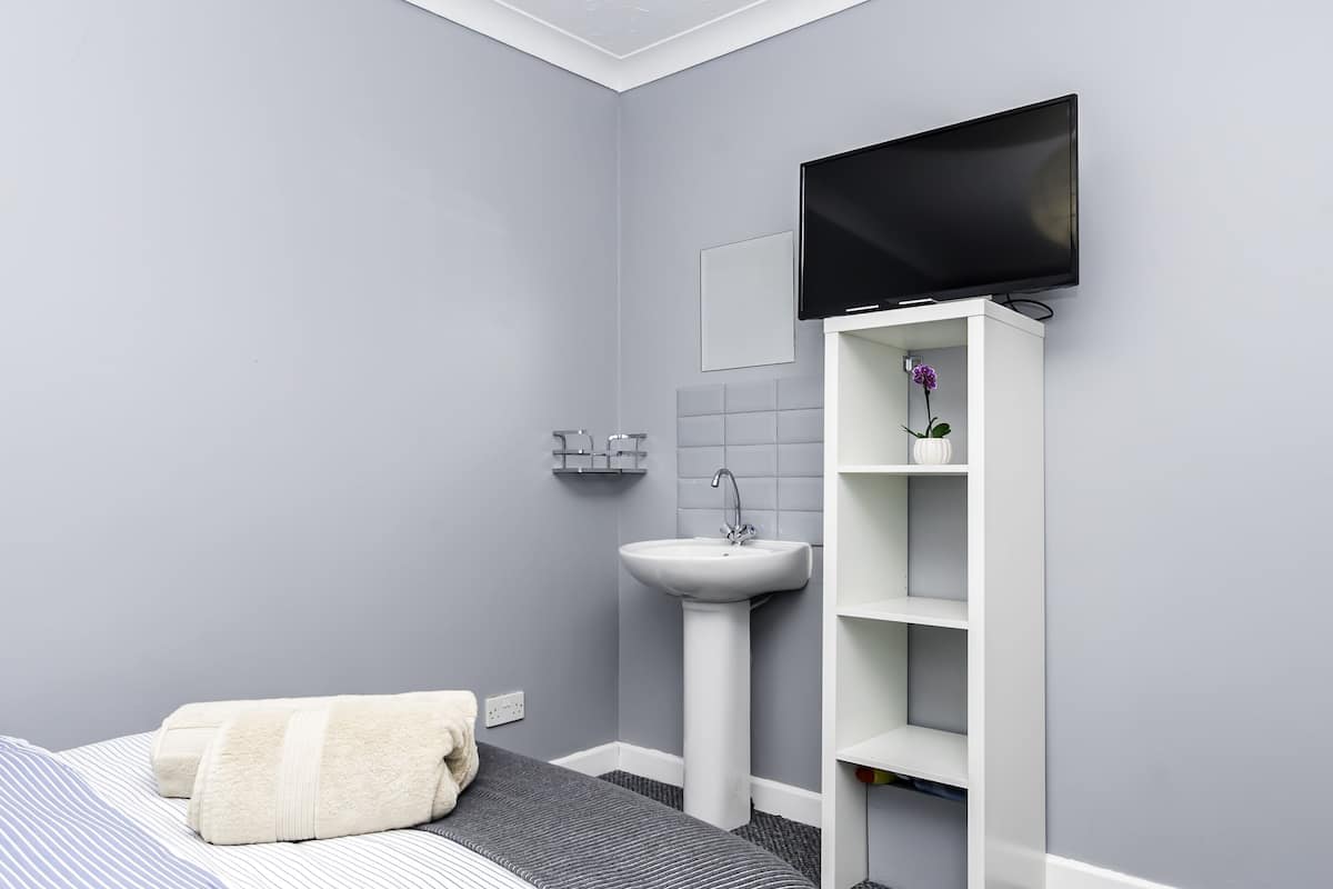 Een moderne badkamer met een grijze wand, een wit wastafelmeubel, een flatscreen-tv bevestigd aan een witte kast en een bed met een gestreept sprei en een opgerolde handdoek.