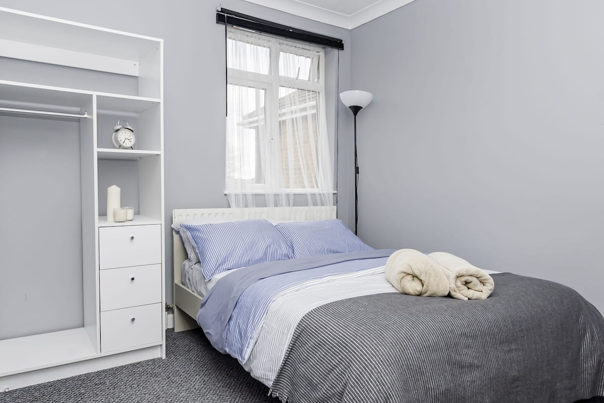 Een slaapkamer met een bed, een kledingkast, een nachtkastje, een lamp en een raam met dunne gordijnen.