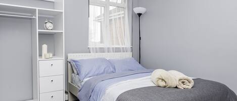 Une chambre à coucher comprenant un lit, une armoire, une table de chevet, une lampe et une fenêtre avec des rideaux légers.