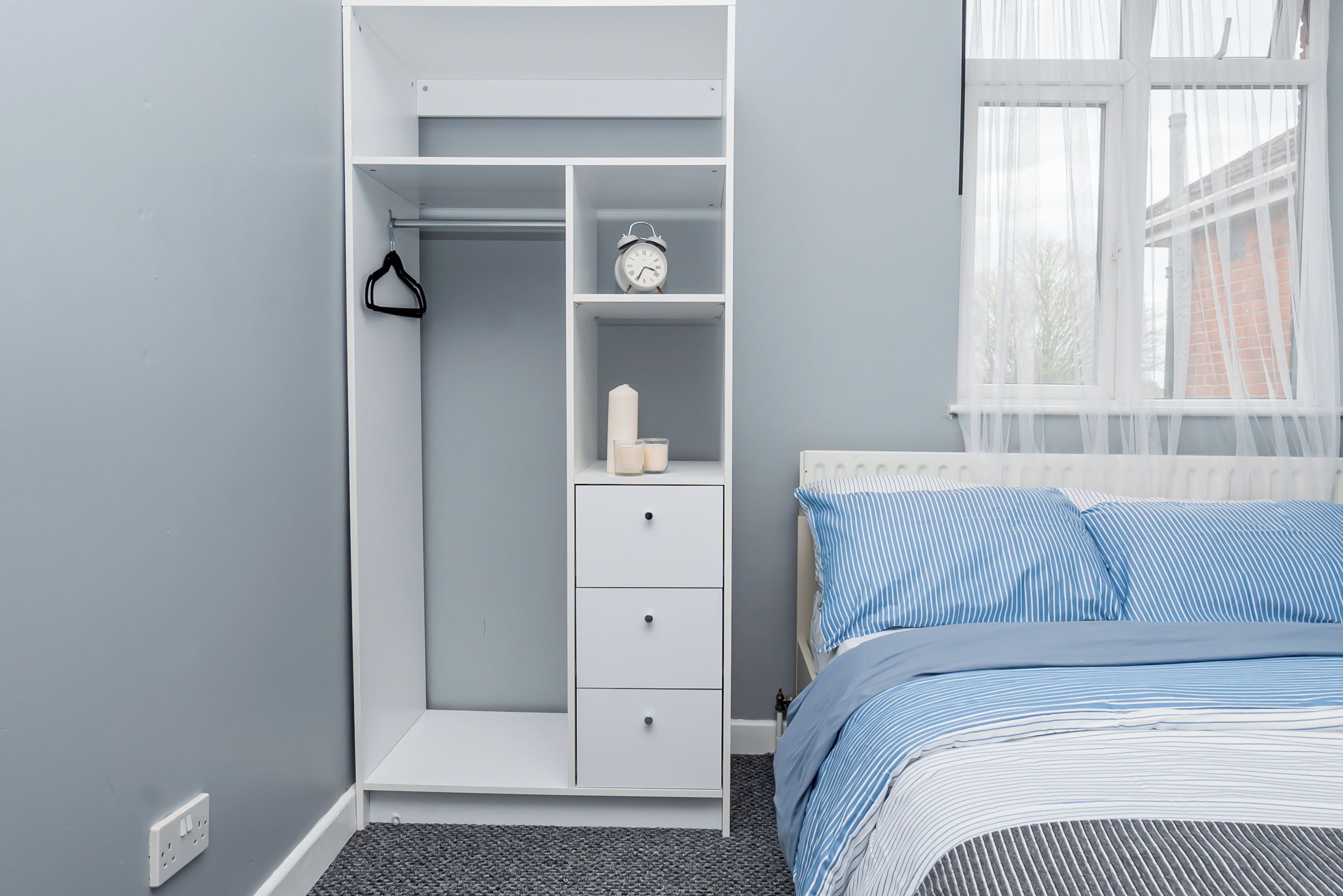 A bedroom with a built-in wardrobe, a bed with striped bedding, and a window with sheer curtains.