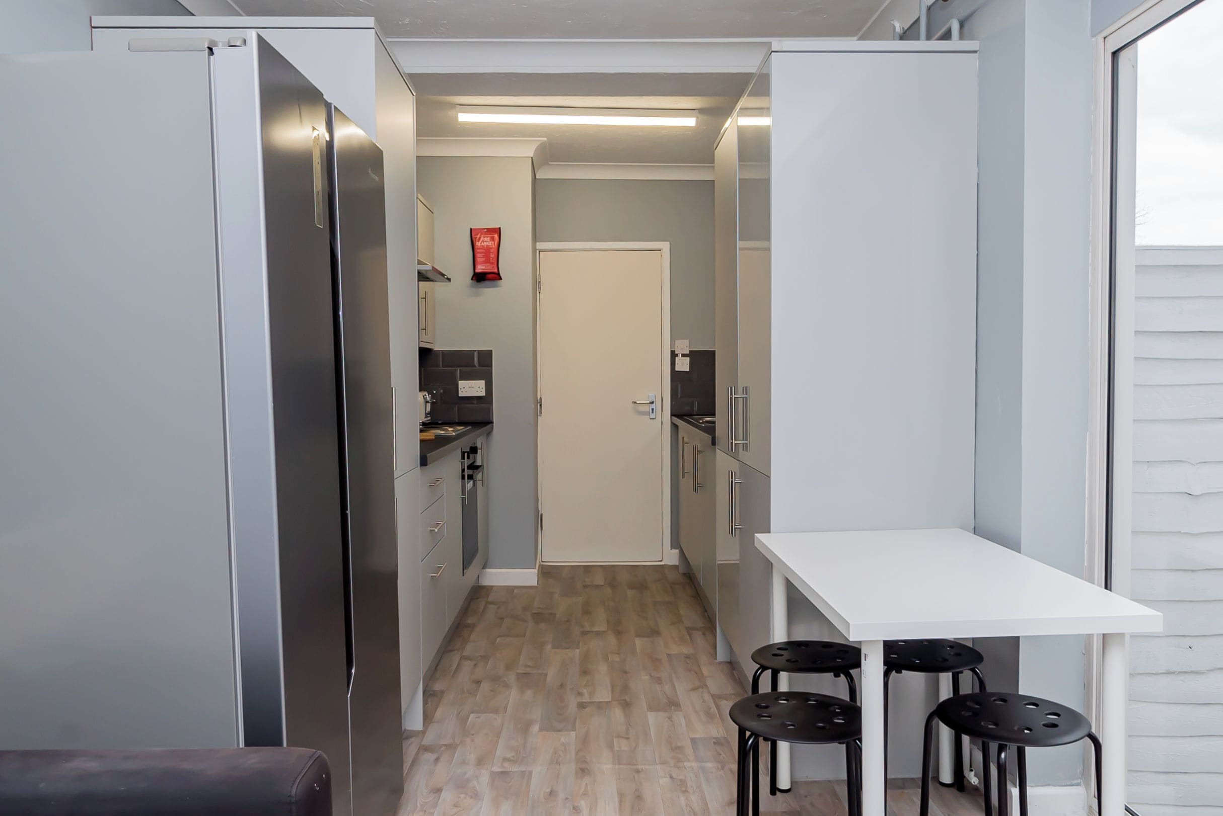 Eine schmale Küche mit Kühlschrank, Schränken und einer weißen Arbeitsplatte. Drei Hocker und ein weißer Tisch sind ebenfalls vorhanden.