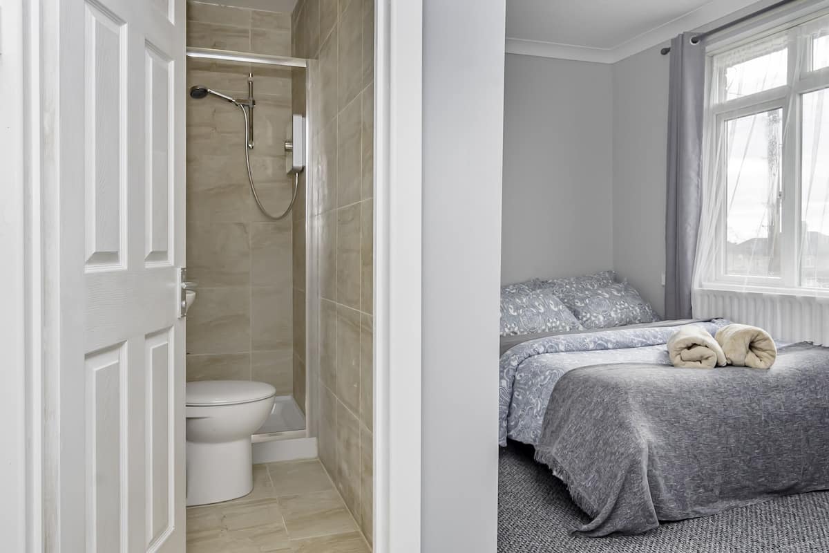 Een badkamer met een douche, toilet en een bed met een grijze sprei en kussens.