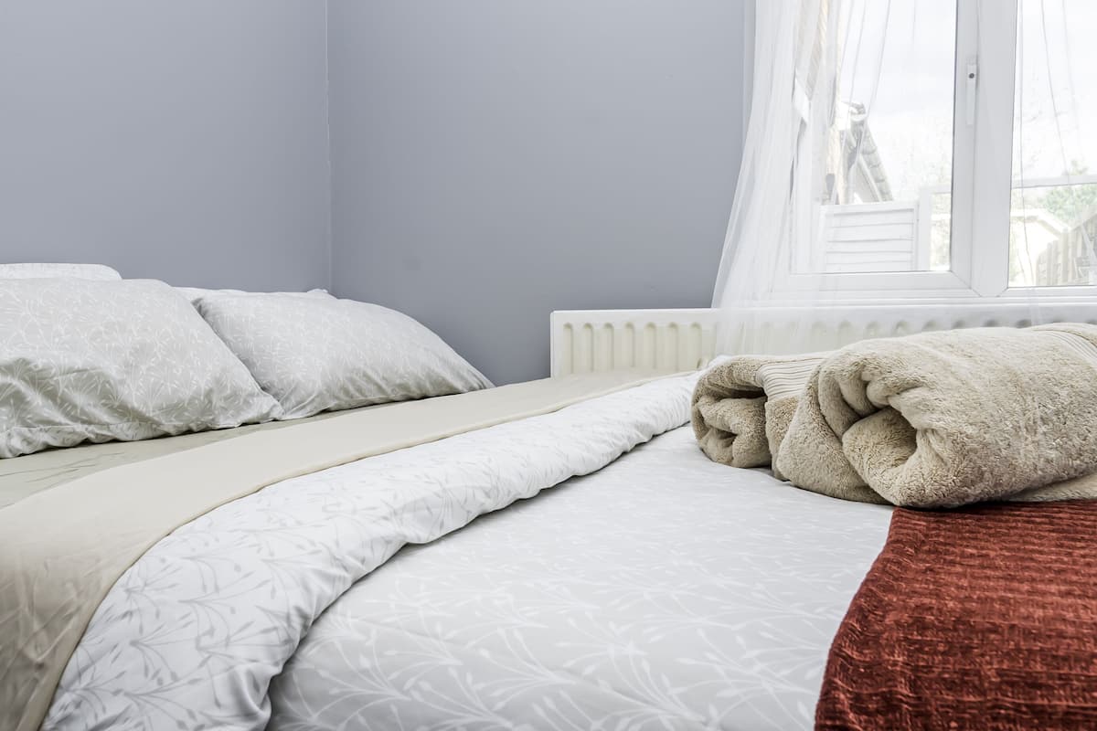 Een net opgemaakt bed met een wit geweven beddekleed en kussens, een opgerolde beige deken, en een raam met dunne gordijnen.