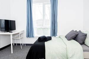 Una habitación de hotel con una cama, un escritorio con televisor, una silla y un ventanal con cortinas.