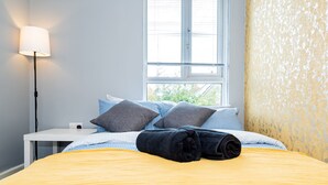 Ein ordentlich bezogenes Bett mit gelber Tagesdecke, zwei Kissen und einer gerollten Decke. Ein weißer Nachttisch mit Lampe und Steckdose ist vorhanden. Ein Fenster mit Jalousien ist sichtbar, und die Wand ist mit Tapete verziert.