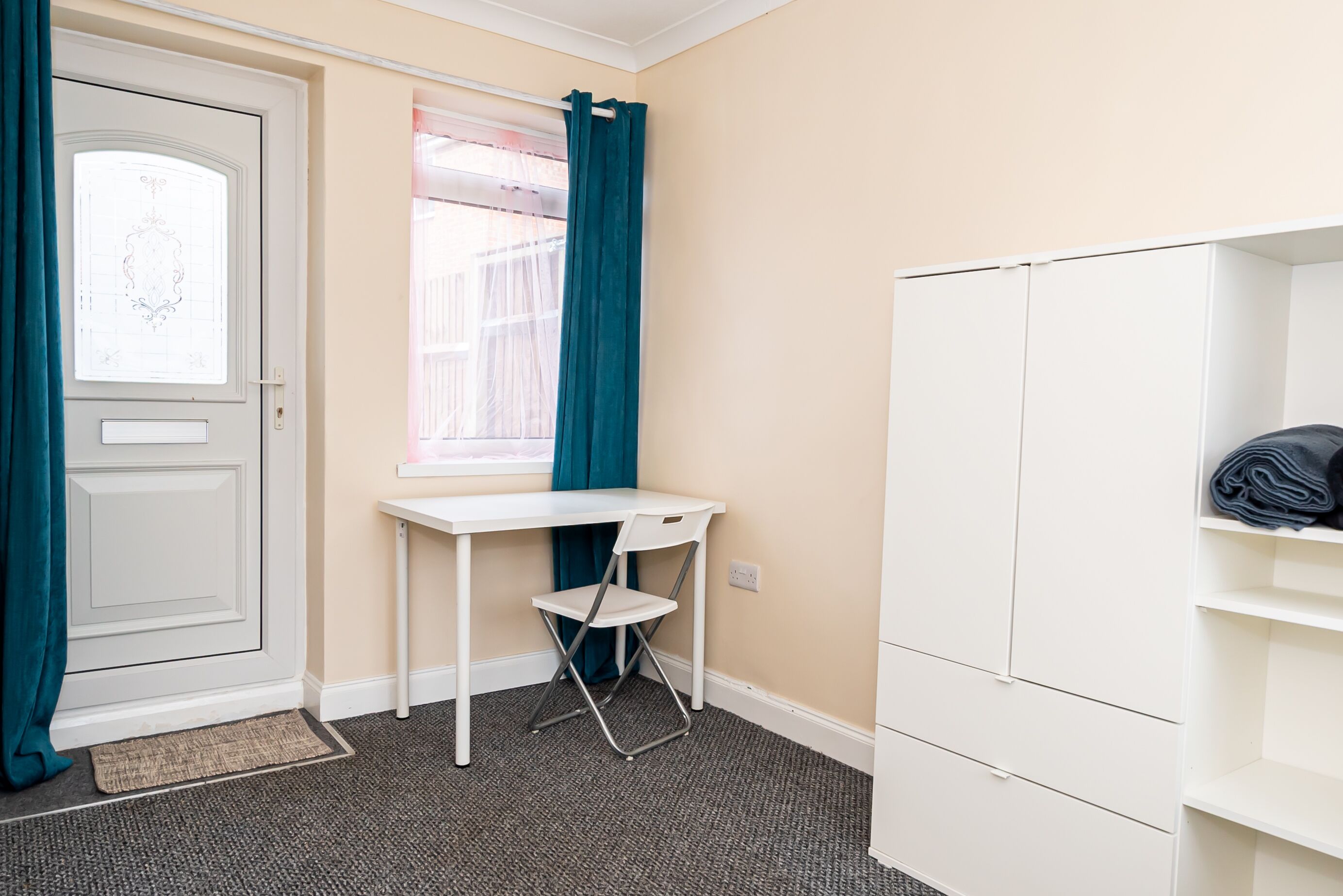 A room with a white door, a window with teal curtains, a white desk, and a white wardrobe.