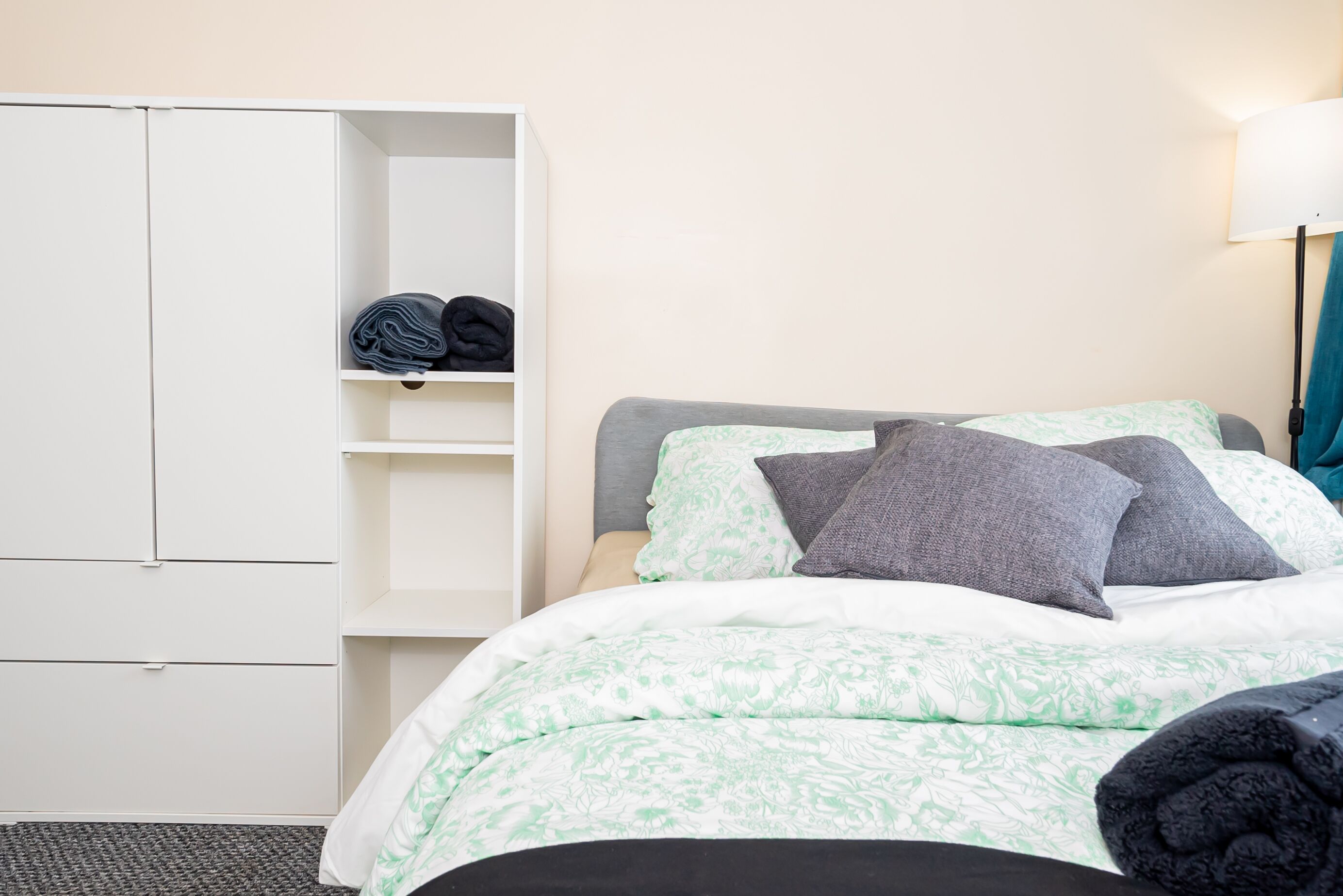 Een net opgemaakt bed met een bloemenprintdeken, een grijze hoofdbord en twee kussens. Links is een witte kast met planken te zien.