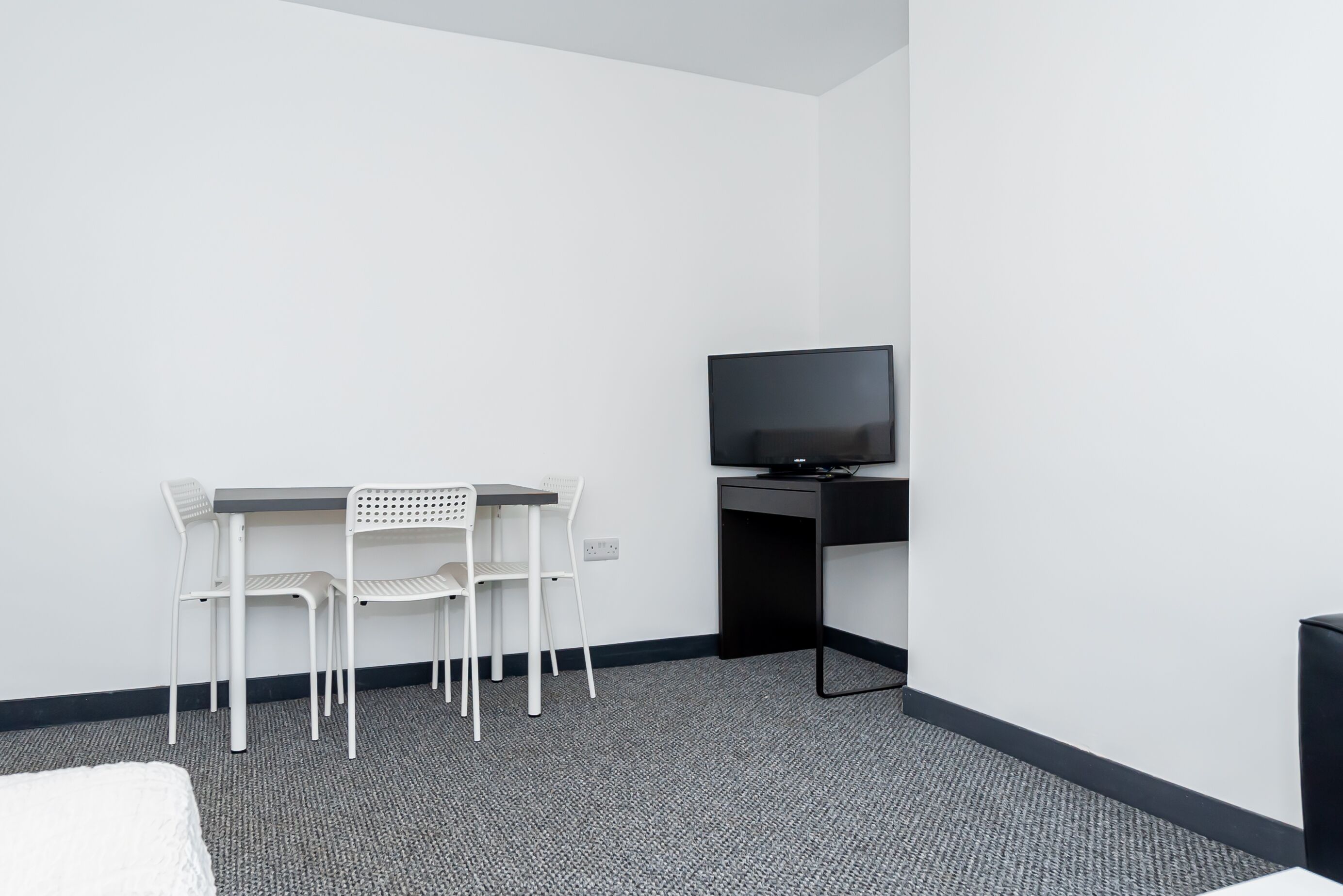 Una habitación de hotel pequeña y moderna con una cama individual, un escritorio con dos sillas blancas, un televisor de pantalla plana y piso alfombrado.