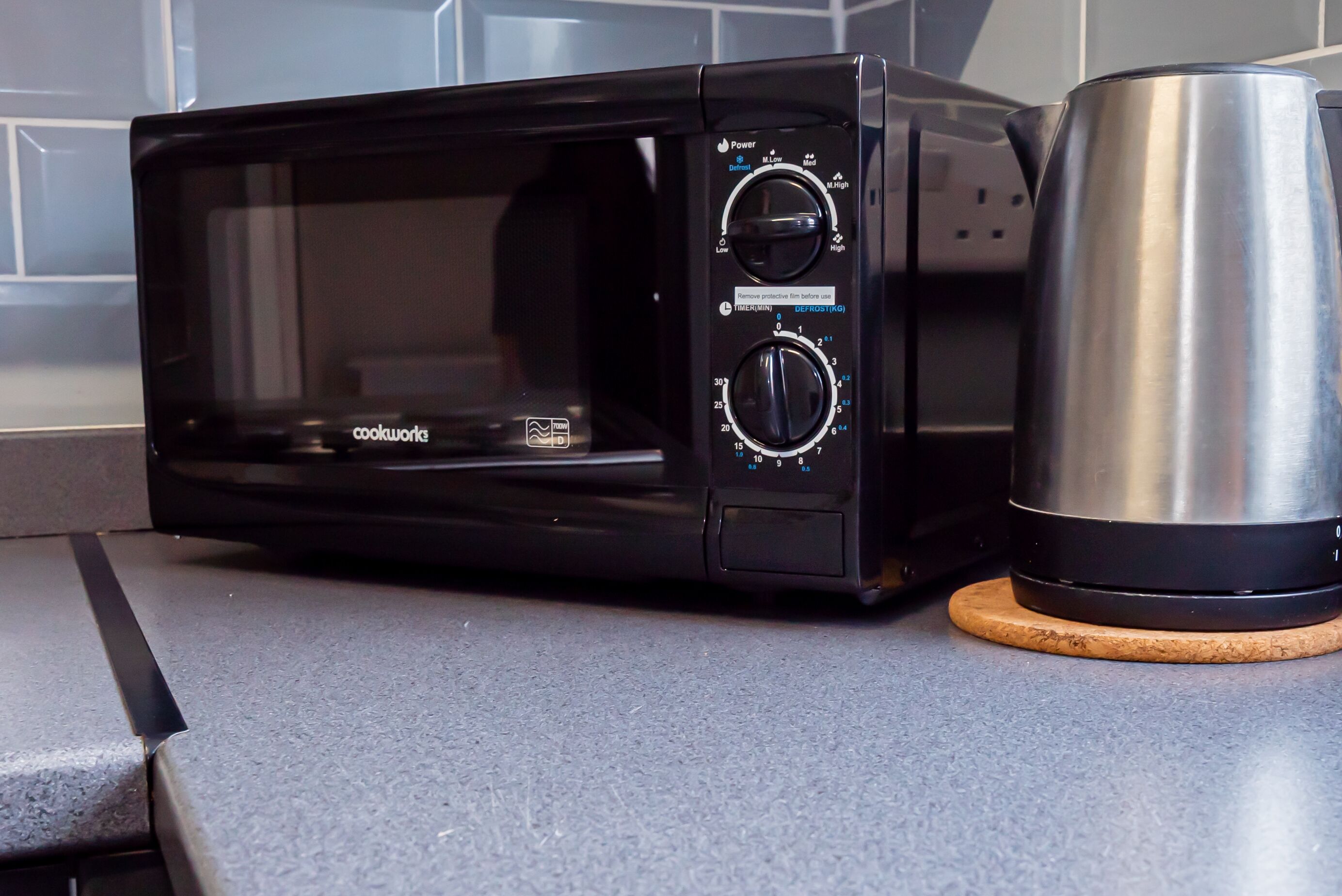 Un horno microondas negro con dos mandos y una pantalla digital, situado sobre una encimera de cocina junto a una cafetera de acero inoxidable.