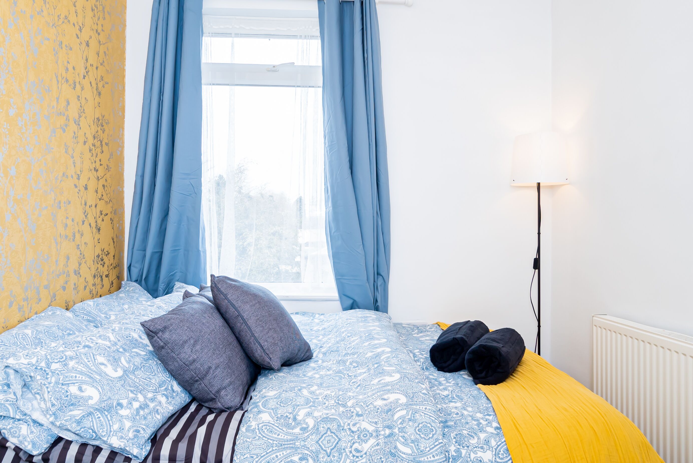 Ein Schlafzimmer mit einem geblümten Bettbezug, zwei Kissen, einem Paar zusammengerollter Handtücher, einer Lampe und einem Fenster mit Vorhängen.