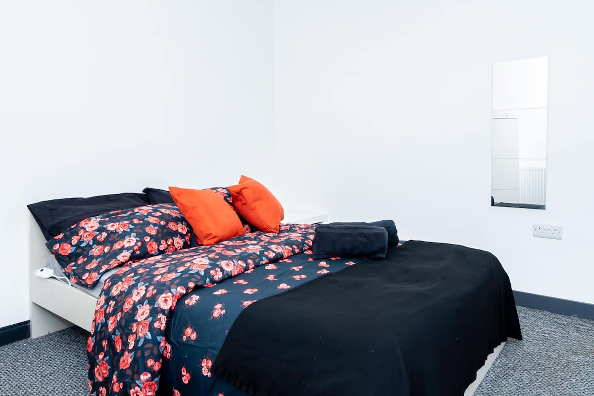 Een net opgemaakt bed met bloemenprint en effen zwarte beddengoed, een witte muur en een raam met jaloezieën.