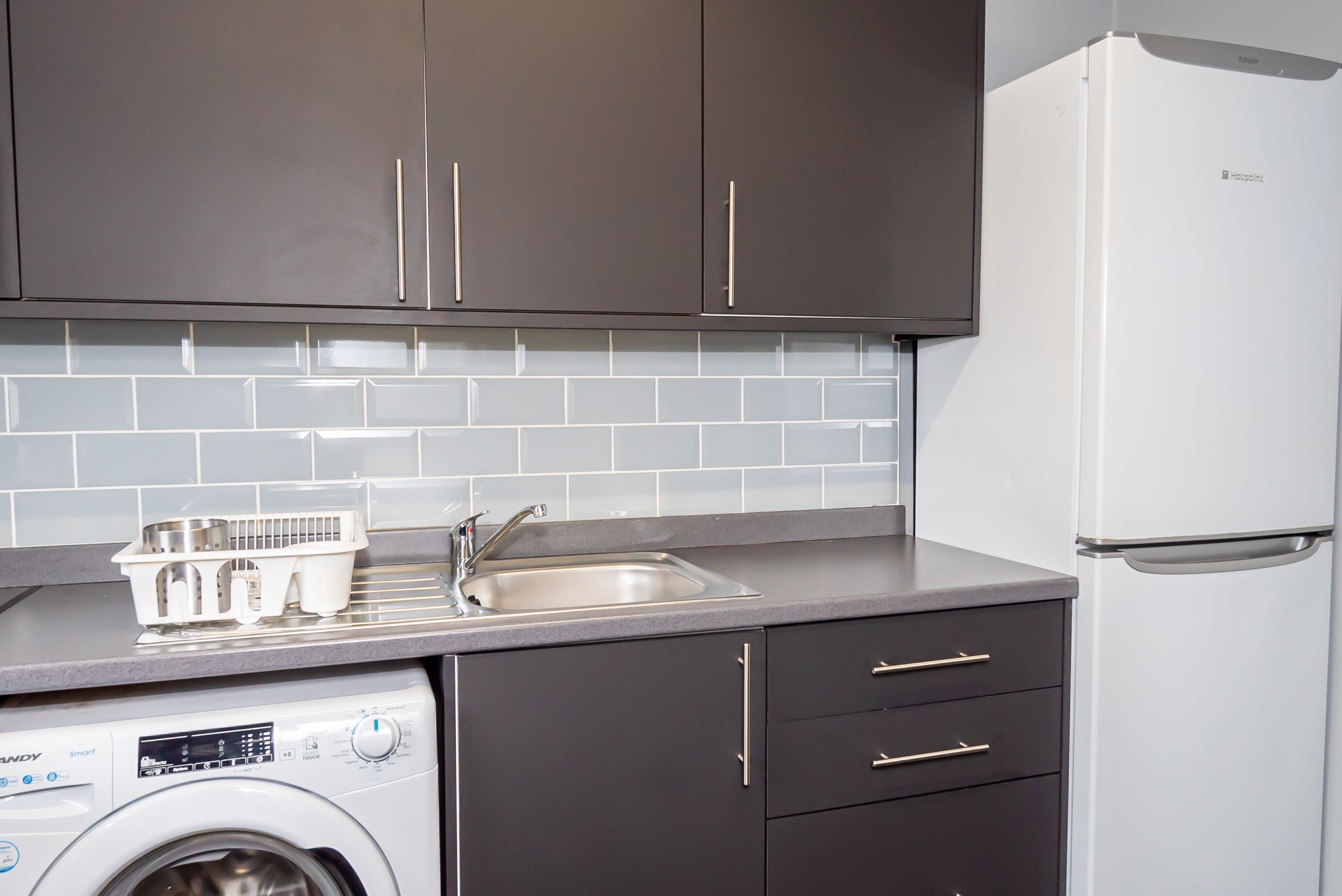 A compact kitchen with dark cabinets, a white washing machine, a stainless steel sink, and a white refrigerator.