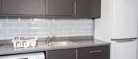 A compact kitchen with dark cabinets, a white washing machine, a stainless steel sink, and a white refrigerator.