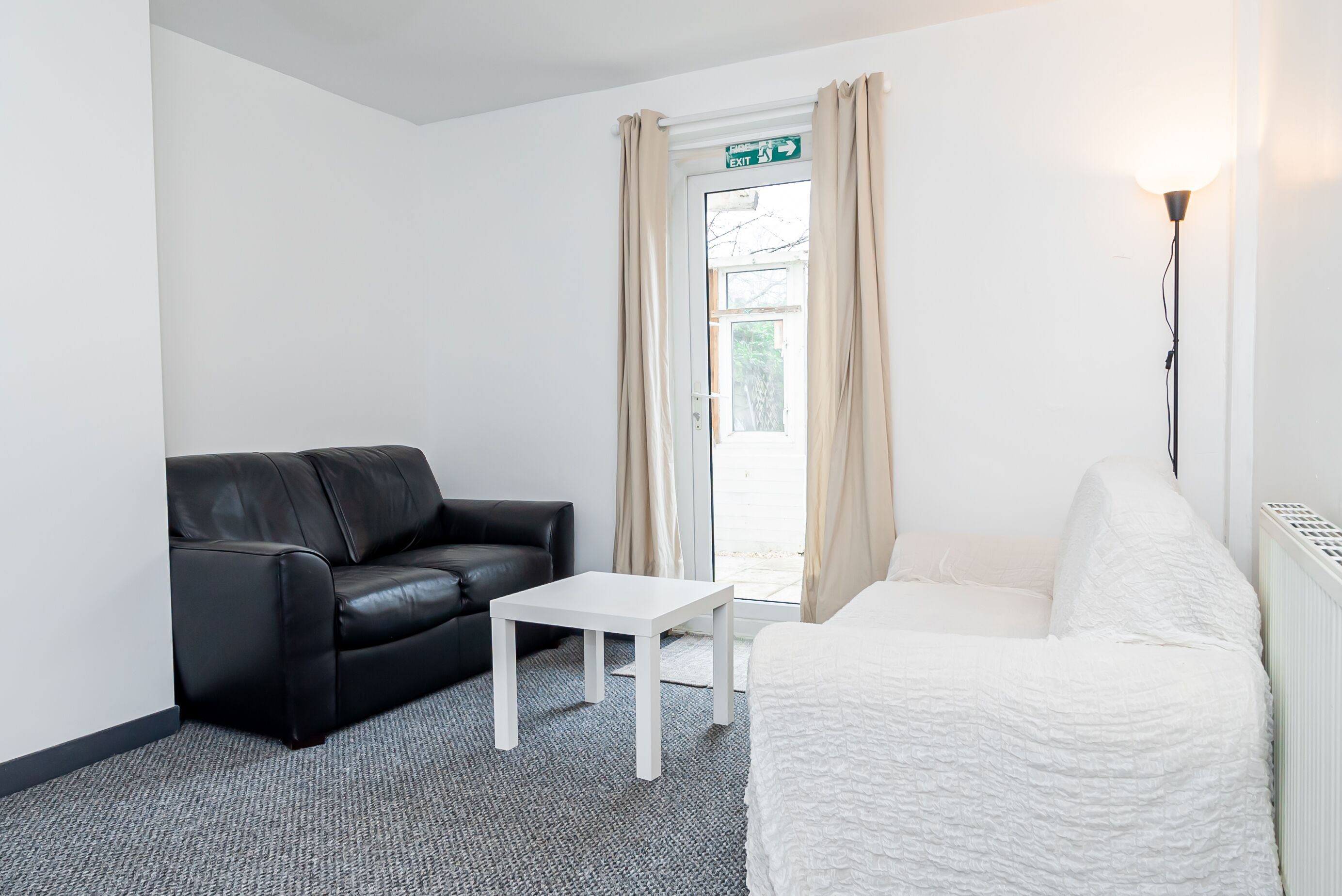 A room with a black leather sofa, a white armchair, a small white table, and a large window with curtains.