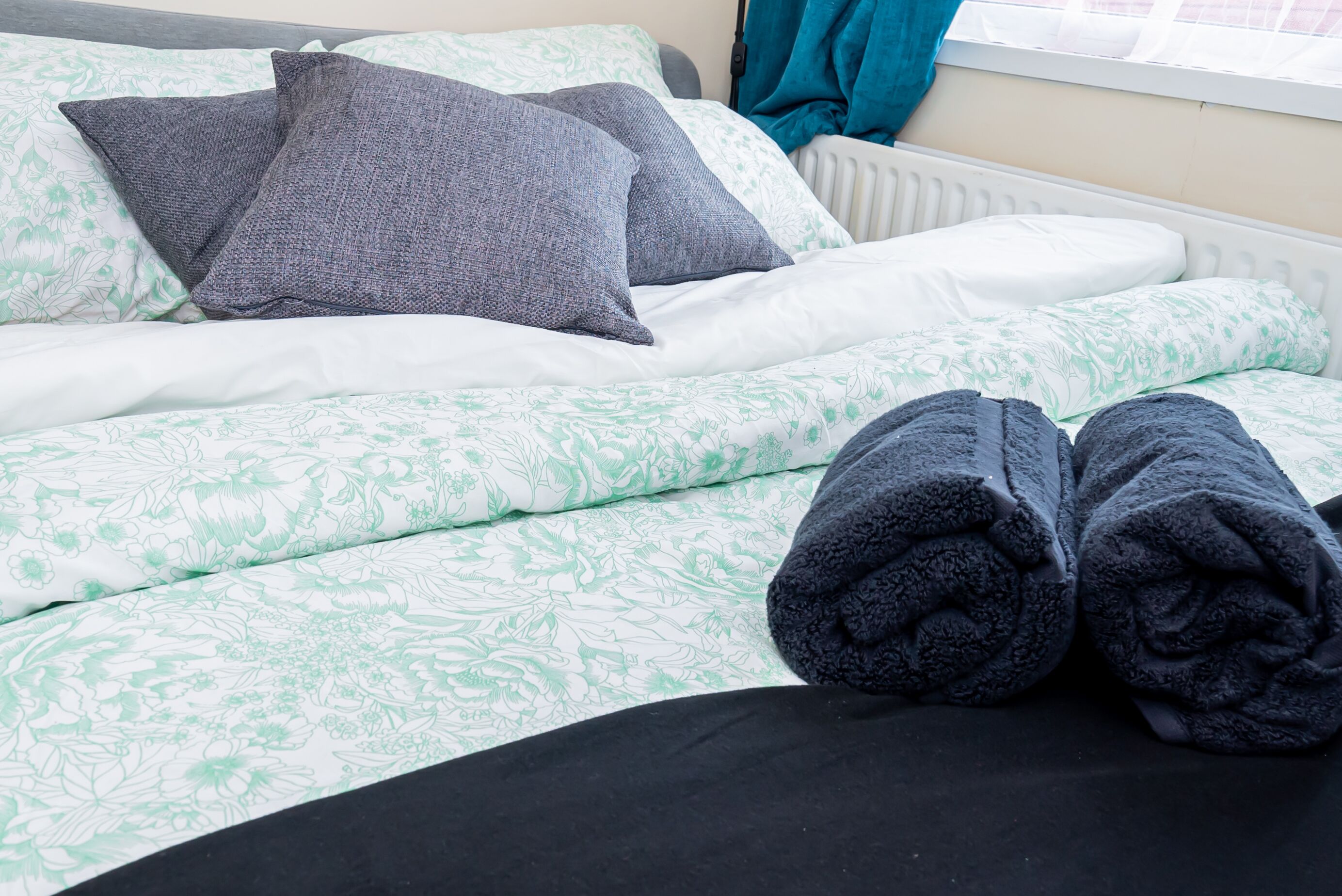 Una cama bien hecha con una colcha de diseño floral, dos almohadas grises y dos toallas enrolladas.
