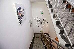 Um corredor com escadas, corrimão, interruptor de luz fixado na parede e porta com placa de saída de emergência verde.