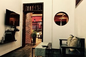 A narrow hallway with a television on the left, a round mirror on the right, and a bench with a cushion.