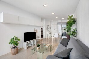 Un salon moderne avec un téléviseur à écran plat, une table basse en verre, un canapé gris et une cuisine en arrière-plan.