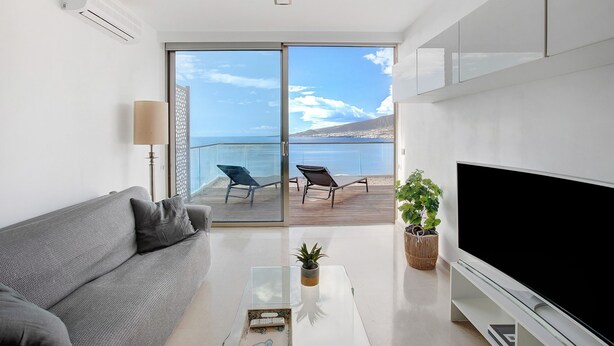Un soggiorno moderno con un divano, una TV a schermo piatto, un tavolino da caffè e un balcone con due sedie vista mare.