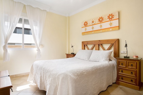 A bedroom with a bed, a nightstand, a dresser, a window with curtains, and a wall-mounted artwork.