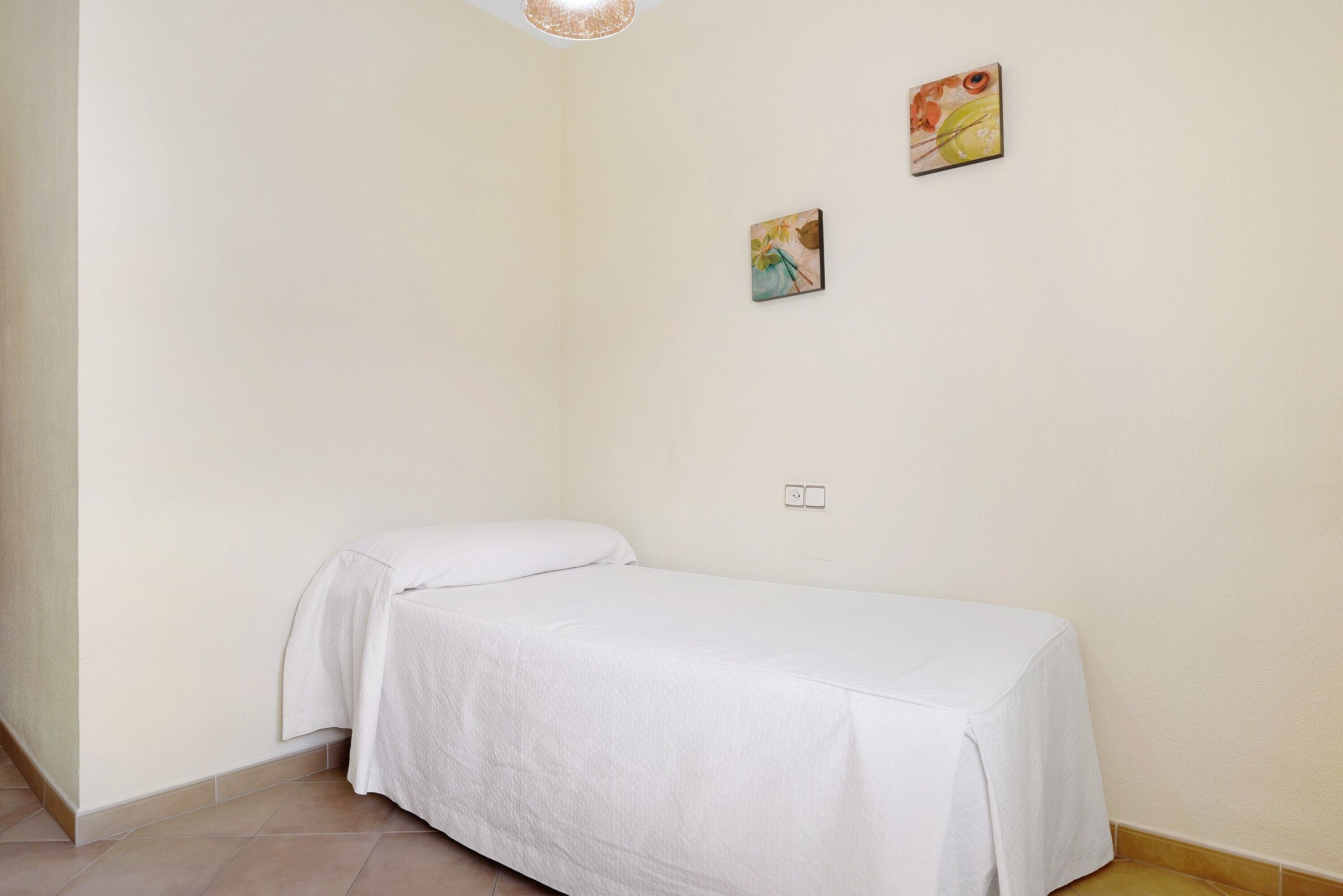 A small, single-bed room with a white bedspread, two small paintings on the wall, and a light fixture above.