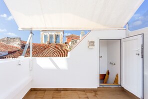 A rooftop terrace with a white awning, a door, and a view of buildings with red-tiled roofs.