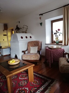 Un salon avec un fauteuil ancien, une petite table sur laquelle se trouve un saladier de fruits, un tapis à motifs et une fenêtre agrémentée d’un vase de fleurs.