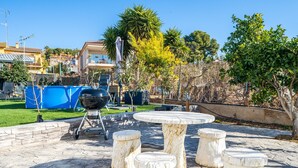 Un espace extérieur aménagé en patio, avec une table et des tabourets en pierre, un barbecue et une clôture bleue en arrière-plan.