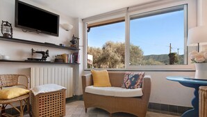 A room with a television, a sofa, a chair, a table, and a window with a view of trees and hills.