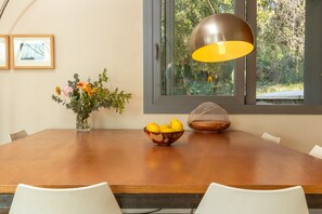 Uma mesa de jantar com cadeiras, um vaso com flores, uma tigela de frutas e uma luminária pendente.