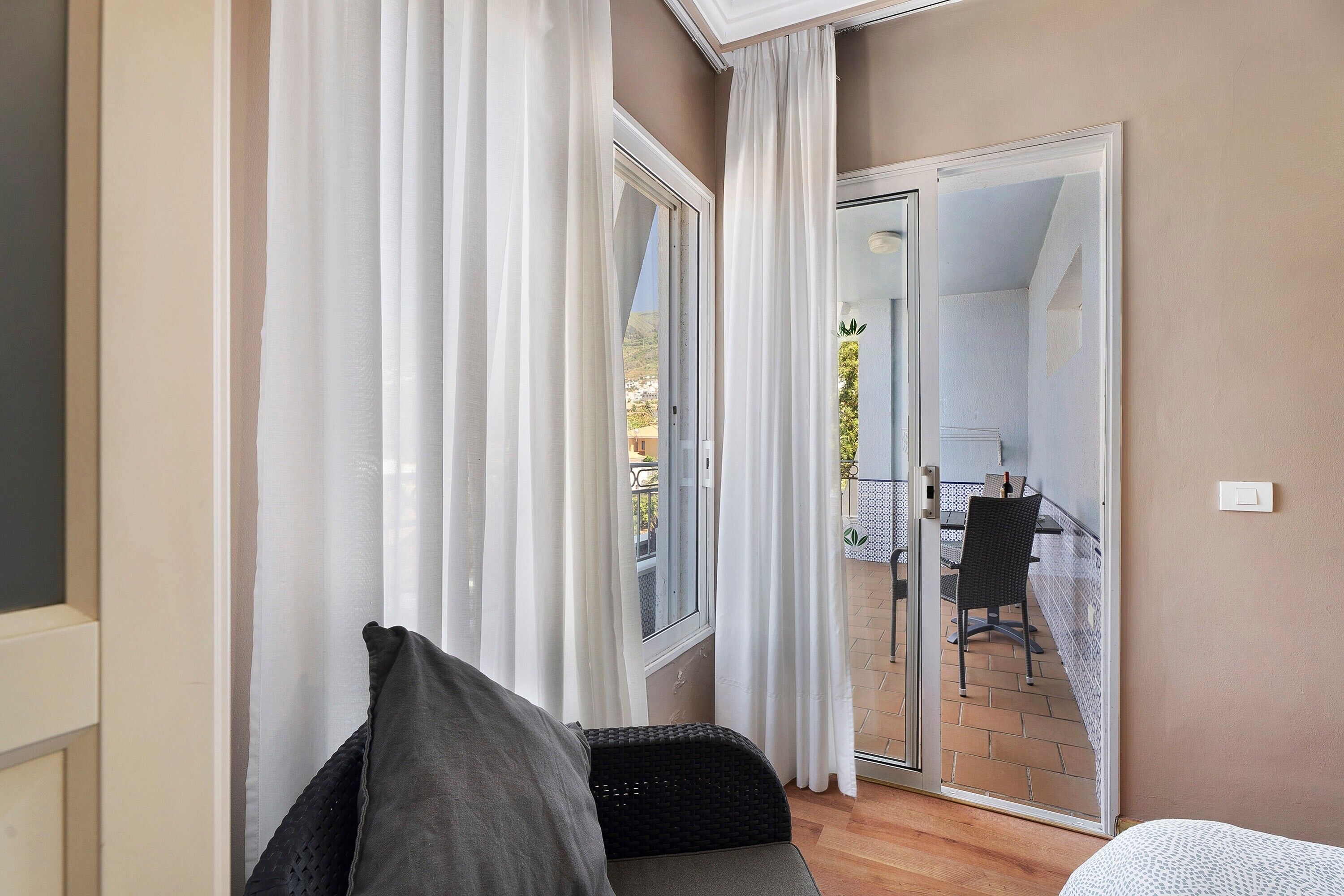 A room with a bed, a window with white curtains, a sliding glass door leading to an outdoor area with a table and chairs.